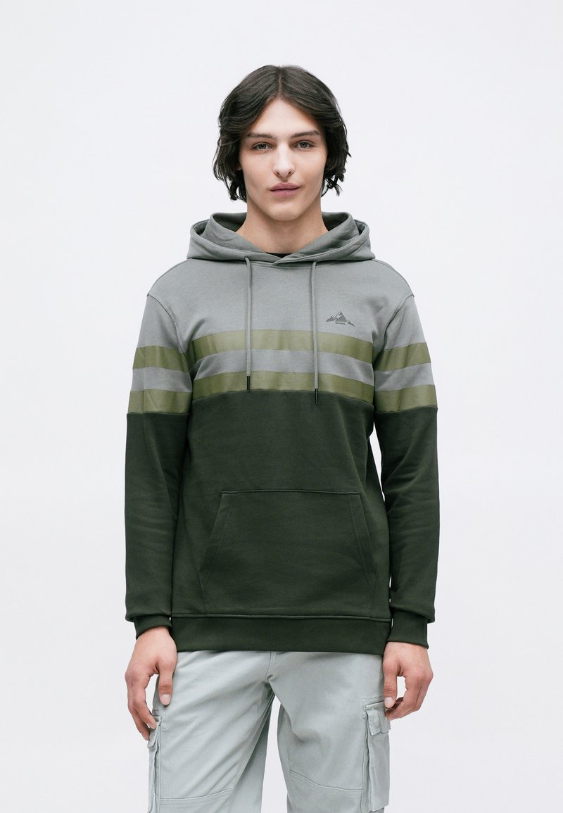 Jeune homme portant un hoodie coloré en vert et gris ainsi que des pantalons cargo gris clair, se tenant devant un fond blanc uni.