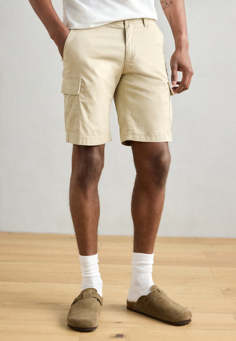 Shorts cargo beiges avec deux poches latérales, fabriqués en tissu de coton. Les jambes se terminent au-dessus du genou, associés à des chaussettes blanches et des sabots marron.