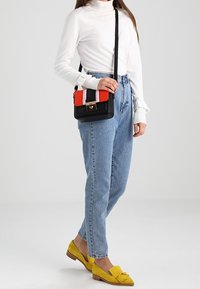 Donna con dolcevita bianco, jeans a vita alta blu, mocassini gialli e che tiene in mano una borsa a tracolla nera, rossa e a righe.