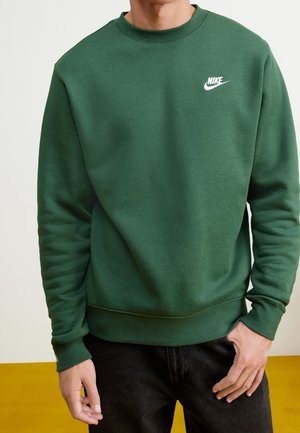 Personne portant un sweat à capuche Nike vert foncé avec un logo blanc sur la poitrine et un pantalon noir, debout avec une main dans la poche.