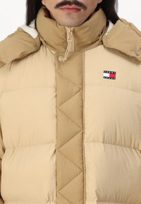 Beige puffjakke med høy krage, quiltet mønster, glidelås og logo-patch. Har en jevn tekstur med kontrasterende detaljer.