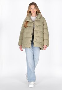 Giovane donna che cammina, indossando un piumino verde chiaro, una maglietta bianca, jeans azzurri chiari e sneakers bianche con plateau, su uno sfondo bianco puro.