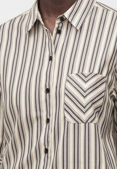 Gros plan d'une personne portant une chemise crème à rayures noires verticales, des boutons noirs et une poche poitrine avec des rayures en chevron.