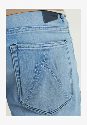 Gaiši zili džinsi ar unikālu dubulto kabatu dizainu aizmugurē, akcentēti ar kontrastējošiem šuvumu un melnu etiķeti.