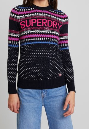 Woman wearing a navy blue sweater with pink, white, and blue patterns and "SUPERDRY" text, paired with light blue jeans.