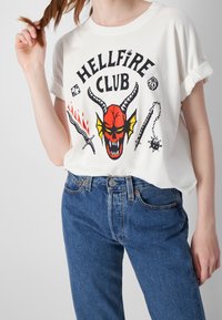 Camiseta blanca con un estampado gráfico colorido que presenta un personaje de diablillo rojo, combinada con jeans de mezclilla azul de cintura alta.