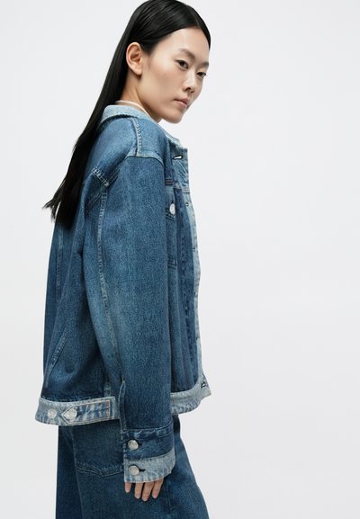 Casaco de ganga azul escuro com um corte ligeiramente oversize. Apresenta uma gola azul clara, botões de metal e costuras contrastantes ao longo das costuras.