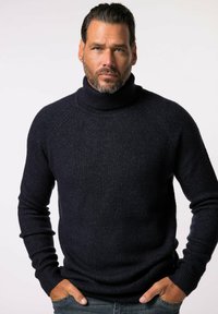 Maglione a collo alto di un blu navy scuro, realizzato in un tessuto a maglia testurizzato, con maniche lunghe e un design aderente, indossato sopra dei jeans di denim.