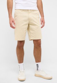 Shorts en coton beige avec une coupe droite, dotés de poches latérales et d'une fermeture à bouton. Associés à des baskets blanches et des chaussettes à motifs.