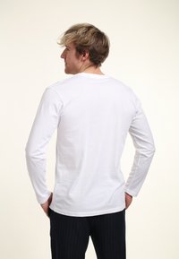 Bílé tričko s dlouhým rukávem z měkké bavlny, s kulatým výstřihem, nošené s černými kalhotami s jemnými pruhy, zachycující pohled zezadu.