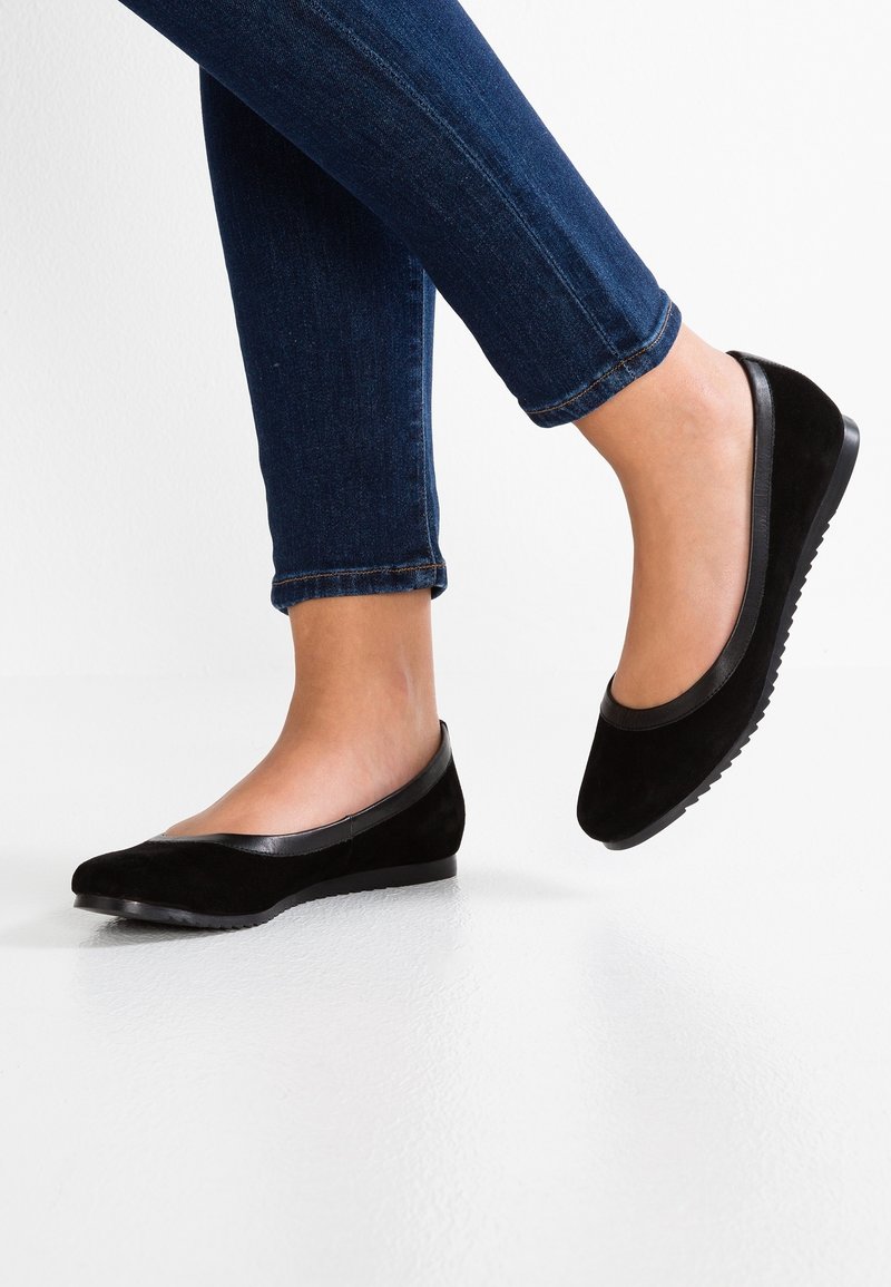 Chaussures plates en daim noir avec une bordure en cuir lisse, bout arrondi et semelle en caoutchouc basse, posées sur un fond blanc avec un pantalon en denim bleu.