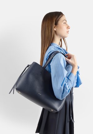 Donna con lunghi capelli castani che indossa una blusa a righe blu e una gonna nera porta una grande borsa tote in pelle blu scuro sulla spalla.
