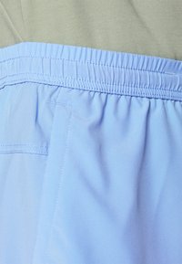 Ceinture élastique bleu clair avec une texture plissée, associée à un tissu gris au-dessus. Un détail de couture plate est visible sur la ceinture.