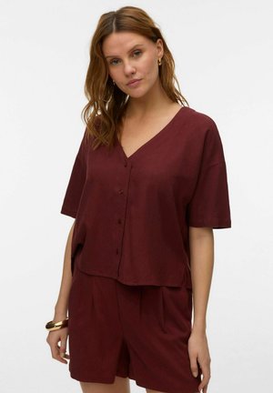 Femme aux cheveux ondulés portant des boucles d'oreilles créoles en or, un bracelet en or, et un ensemble assorti de chemise à boutons et short bordeaux à col en V.