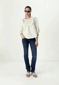 Mujer con cabello trenzado que lleva gafas de sol oscuras, blusa blanca de tela de ojal con mangas abullonadas, jeans azul oscuro y sandalias de tacón blancas, de pie de manera relajada.