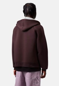 Sudadera marrón con un ajuste holgado, capucha oversized y puños acanalados. Combinada con pantalones cargo de color púrpura claro que cuentan con bolsillos.