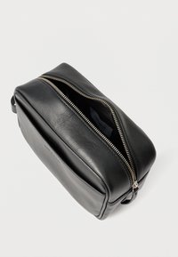 Crna kozmetička torbica od kože s glatkom teksturom, gornjim zatvaračem na patent i bočnom ručkom. Ima jedan odjeljak s tkaninskom podlogom.