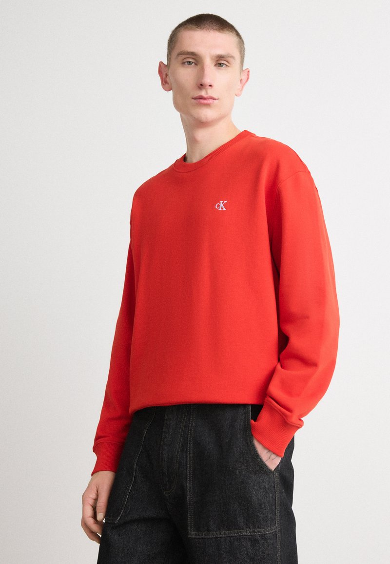 Κόκκινη ζακέτα crewneck από μείγμα βαμβακιού, με ραμμένες μανσέτες και ένα μικρό κεντητό λογότυπο στο στήθος; συνδυασμένη με σκούρα τζιν παντελόνια.