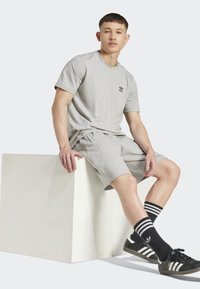 Gray cotton t-shirt and shorts, sitting on a white cube. Black Adidas logo, striped black and white socks, and black sneakers with white accents.