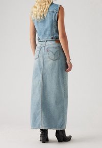 Lange Jeans-Maxikleid in Hellblau, mit ärmellosem Crop-Top, langem Rock und Rückentaschen. Der Saum hat einen fransigen Rand. Getragen mit schwarzen, hohen Stiefeln.