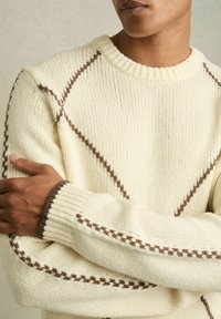 Krēmkrāsas adīts džemperis ar brūniem diagonāliem šuvju akcentiem, ribota apaļa kakla izgriezuma un aprocēm. Tekstūra izskatās maiga un masīva.