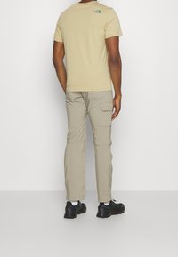 Camiseta beige con un logotipo verde en la parte trasera, combinada con pantalones cargo de color caqui claro que cuentan con bolsillos laterales y zapatillas deportivas negras.