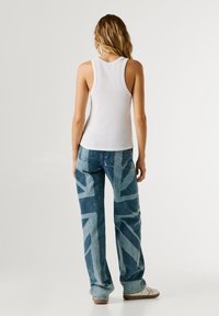 Achteraanzicht van een persoon die een wit geribbeld tanktop en blauwe spijkerbroek draagt met een gedurfd wit geometrisch patroon en lichtgekleurde stiksels.