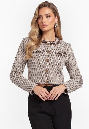 Donna con lunghi capelli biondi mossi, che indossa una giacca corta beige e marrone a motivo con bottoni dorati e pantaloni neri, guarda di lato.