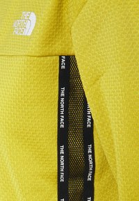 Tissu texturé jaune avec maille noire et bande noire étiquetée "THE NORTH FACE" en texte blanc.