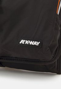 Schwarze K-Way-Tasche aus einem glatten, glänzenden Stoff, rechteckiger Form und einem auffälligen weißen Logo-Tag mit schwarzem Rand. Akzente setzen orangefarbene Nähte.