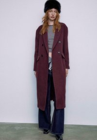 Burgundy dobbeltradet frakke med store sorte knapper, båret over en stribet top og parret med vidde ben i denim og sorte hæle.