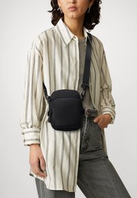 Borsa a tracolla nera realizzata in materiale liscio, di forma rettangolare, con zip e tracolla regolabile, indossata sopra una maglietta a righe.