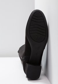 Bottes en daim noir avec talon bloc, positionnées semelle vers le haut montrant la taille 37 et le nom de la marque Unisa sur une surface et un mur blancs.