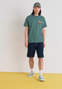 Camiseta de algodón verde con un gráfico colorido en el pecho, combinada con shorts de mezclilla oscura. Zapatillas azul claro y gorra verde completan el look.