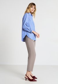 Donna che indossa una camicia a righe blu a maniche lunghe, pantaloni beige dritti e décolleté borgogna con punta, con fiocchi, in piedi di lato.