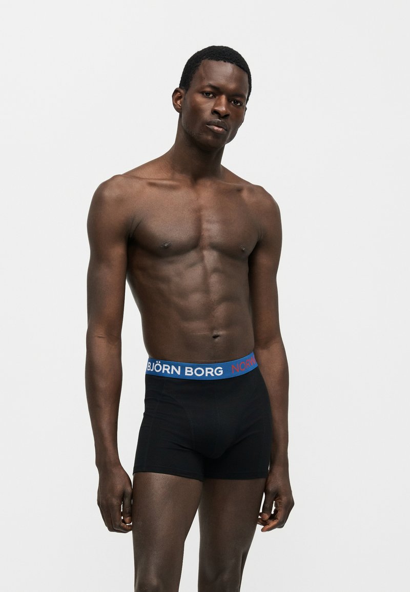 Μυώδης άνδρας που στέκεται μπροστά σε απλό φόντο, φοράει μαύρα μπόξερ με μπλε ζώνη που αναγράφει «Björn Borg».