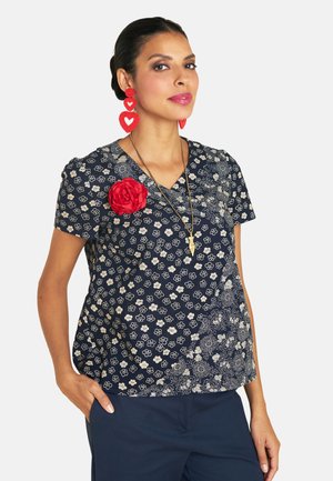 Marineblaue Bluse mit floralem Muster und kurzen Ärmeln. Sie hat ein rotes Stoffblumenakzent und einen V-Ausschnitt, kombiniert mit dunklen Hosen.