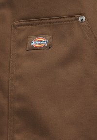 Καφέ ύφασμα με ραμμένη μπροστινή τσέπη που φέρει ένα μικρό ταμπελάκι με το λογότυπο Dickies και μεταλλικό καρφί στο πάνω γωνιακό σημείο.