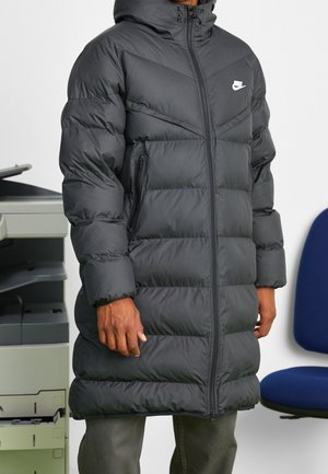 Osoba ubrana w długą czarną kurtkę puchową Nike z kapturem, stojąca obok drukarki biurowej i niebieskiego krzesła.