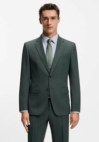 Dunkelgrüner, maßgeschneiderter Anzug aus strukturiertem Stoff mit Reverskragen, schmal geschnittenen Hosen, einem hellblauen Hemd und einer gemusterten Krawatte.