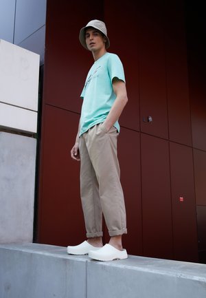 Hellgrünes kurzärmliges Hemd mit Grafikdruck, beiger Baggy-Hose und weißen Slippern. Grauer Bucket Hat. Urbane Umgebung.
