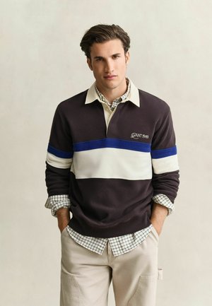Junger Mann mit Händen in den Taschen, trägt ein dunkles Rugbyshirt mit weißen und blauen Streifen über einem karierten Hemd und beigen Hosen.