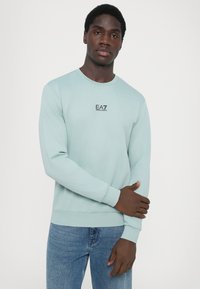 Pulover crewneck de culoare verde menta deschis, cu mâneci lungi, având un mic logo negru EA7 pe piept. Material și croi confortabile.