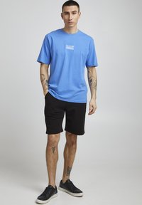 Tricou din bumbac albastru cu un mic design grafic alb pe piept, asociat cu pantaloni scurți negri și adidași negri. Tatuaje vizibile pe brațe.