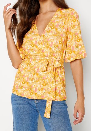 Vrouw draagt een gele bloemenwikkeltop met korte pofmouwen en een gestrikte taille, gecombineerd met een blauwe spijkerbroek, poserend met één hand bij haar haar.