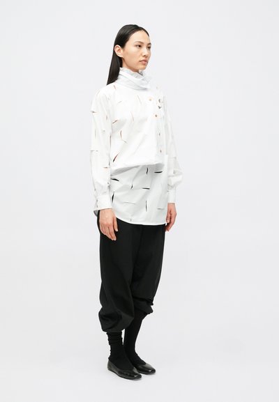 Λευκό πουκάμισο με κουμπιά, διακοσμημένο με αφηρημένα μαύρα και πορτοκαλί μοτίβα, συνδυασμένο με φαρδιά μαύρη παντελόνια και μαύρα παπούτσια με επίπεδη σόλα.