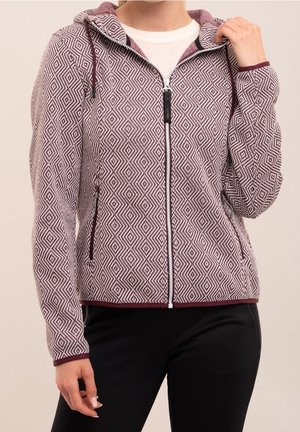 Femme portant un sweat à capuche zippé à motif géométrique bordeaux et blanc avec une capuche et un pantalon noir, tenant le col du sweat à capuche d'une main.