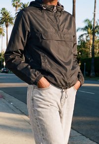 Personne portant une veste coupe-vent noire à capuche et un jean gris clair, les mains dans les poches, debout dans une rue ensoleillée avec des palmiers.