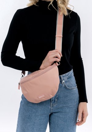 Kobieta w czarnej bluzce z długim rękawem i niebieskich dżinsach, trzymająca jasnoróżową torebkę na ramię z szerokim paskiem przewieszonym przez pierś.