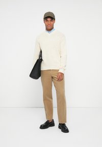 Homem vestindo calças de veludo cotelê bege, suéter de malha creme, camisa azul, boné xadrez, sapatos loafers pretos, segurando uma bolsa tote de couro preta.